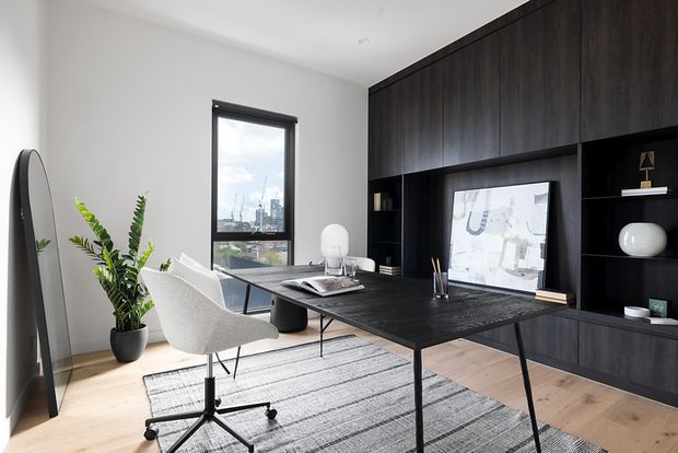 A refined home office with a large black built-in wall of cabinetry and open shelving, a dark desk, a white upholstered swivel chair, and a pale timber floor. A tall window overlooks a city skyline, while a large grey striped rug, potted plant, floor mirror, and minimal decorative objects add warmth and texture.