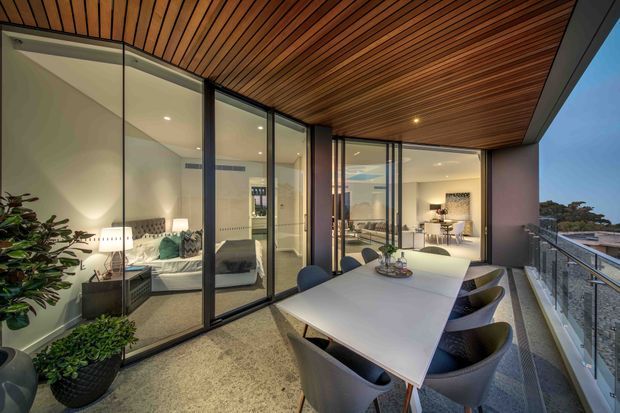 Contemporary covered balcony terrace with a large outdoor dining table and chairs, overlooking the surrounding suburban landscape. Floor-to-ceiling black-framed glazing opens views into a stylish living room and dining area, while a warm timber slatted soffit creates a sheltered, refined outdoor entertaining space. Potted plants and integrated lighting add warmth.