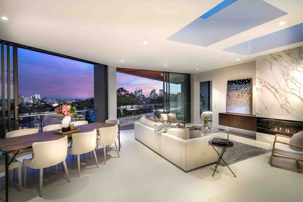 Contemporary open-plan apartment interior combining a dining area and living room, with floor-to-ceiling glazing overlooking a city skyline at dusk. The room features a white sectional sofa, sculptural lounge seating, a dark dining table with upholstered chairs, recessed lighting, a large marble-look feature wall surrounding a fireplace or media unit, and geometric ceiling recesses with blue accents.