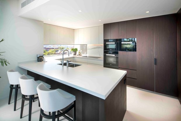 Bright contemporary kitchen featuring a large white waterfall-edge island with integrated sink and three white stools, pale full-height cabinetry, a marble-look splashback, and a contrasting dark timber-toned wall of tall cabinetry with integrated appliances. Recessed ceiling lighting and clean, minimal lines create an airy, refined space.