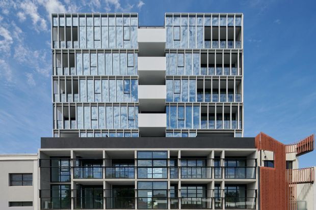 Street-facing photograph of a contemporary multi-storey apartment building with a symmetrical facade, extensive floor-to-ceiling glazing, vertical metal fins, projecting balconies, dark lower-level framing, and a contrasting terracotta-toned screen element. The building is shown against a blue sky.