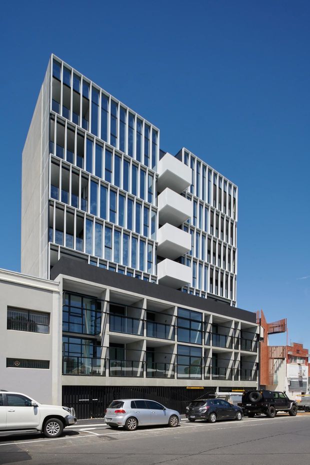 Street-level view of a contemporary multi-storey urban apartment or mixed-use building. The facade combines extensive blue-tinted glazing, repeated vertical window fins, pale concrete or rendered panels, projecting white balconies, and a darker podium with glazed frontage. Adjacent older low-rise commercial buildings and parked vehicles are visible along the street.