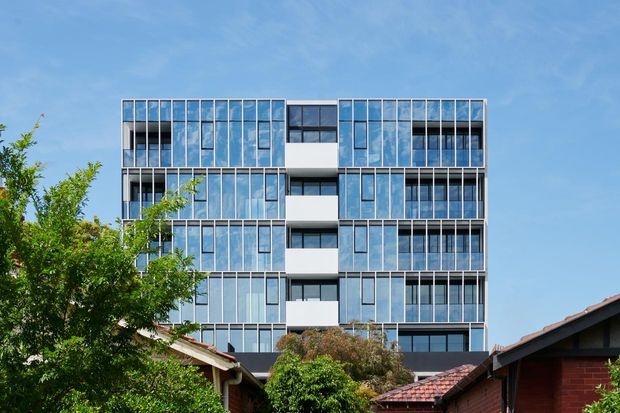 A contemporary multi-storey apartment building viewed from outdoors, featuring a highly regular facade of blue-tinted floor-to-ceiling glazing, slender vertical mullions, dark window bands, and projecting white balcony or wall volumes. Established low-rise residential rooftops and mature trees occupy the foreground beneath a clear blue sky.