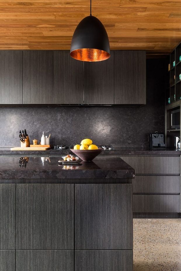 A dark, sophisticated contemporary kitchen with full-height charcoal textured cabinetry, a dark stone backsplash and island benchtop, and warm timber ceiling boards. A large black pendant light with a copper-toned interior hangs above the island, while integrated appliances and open shelving occupy the right side. The kitchen is styled with a fruit bowl and small countertop accessories, and a terrazzo-like floor adds contrast.
