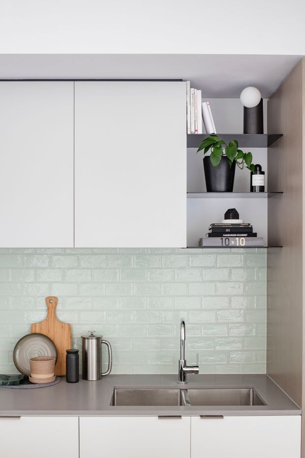 A compact, contemporary kitchen vignette with white flat-panel overhead and base cabinetry, open black shelving, pale mint-green glossy subway-tile splashback, and a grey benchtop. A double stainless-steel sink with a gooseneck tap sits beneath the shelving, alongside a timber chopping board, cookware, and small decorative objects. The cabinetry extends to the ceiling, creating a clean, space-efficient composition.