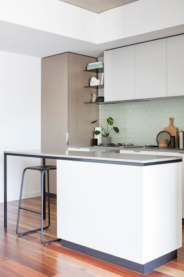 A bright, compact contemporary kitchen featuring a white island with dark edging and base trim, integrated white overhead cabinetry, pale timber-toned tall cabinetry, and open black shelving. The island includes a slim black-framed breakfast bar with two minimalist stools. A pale green glazed brick backsplash, stainless-steel cooking appliances, potted plant, and timber serving boards add texture and colour. Warm timber flooring and a recessed concrete ceiling feature complete the clean modern composition.