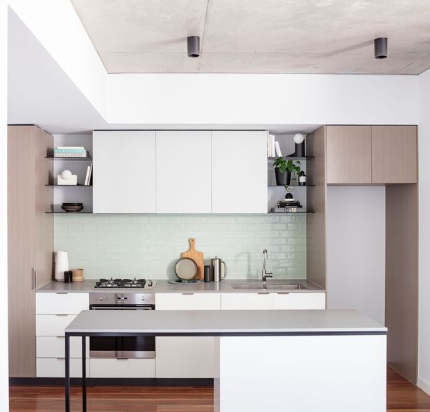 A bright, compact contemporary kitchen with a white island bench, pale timber-toned tall cabinetry, white overhead cabinets, open shelving, and a pale green glazed brick backsplash. Integrated appliances, a gas cooktop, sink, and timber flooring are visible. The ceiling has a raw concrete finish with black cylindrical downlights, creating a restrained modern aesthetic.