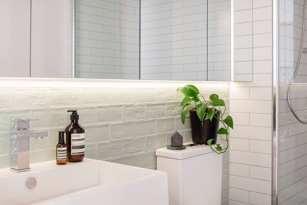 Bright, compact contemporary bathroom featuring a white wall-mounted vanity with chrome tapware, a white bathtub or basin in the foreground, pale green textured subway-tile splashback, white subway-tiled shower walls, mirrored cabinetry, and a glass shower enclosure. A potted plant and toiletry bottles add soft decorative detail.