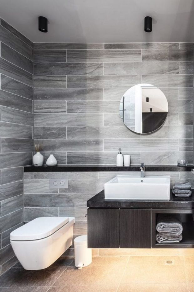 A contemporary bathroom with grey wood-look wall tiles arranged in a horizontal staggered layout, a dark floating vanity with a white vessel-style basin, wall-mounted toilet, round mirror, black shelf, and minimalist black ceiling spotlights. White accessories and neatly folded towels add contrast, while the warm grey tiled floor creates a clean, refined finish.