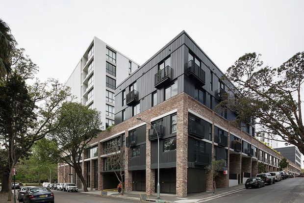 Street-level exterior view of a contemporary multi-storey apartment building. The composition combines a lower and mid-level facade in exposed brown brick with dark metal-clad upper volumes, projecting balconies, extensive dark-framed glazing, and a taller white apartment wing behind. The building fronts an urban street lined with mature trees and parked vehicles.