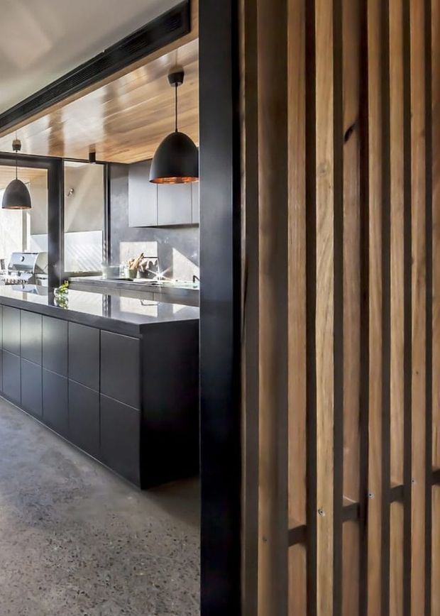 A contemporary kitchen viewed through a vertical timber-slat divider. The space features a dark island with a reflective stone benchtop, matte dark cabinetry, a warm timber-lined ceiling, black pendant lights, concrete flooring, and a pale stone or concrete backsplash. Black-framed glazing and strong sunlight add contrast, while the slatted timber screen creates a layered entry or partition element.