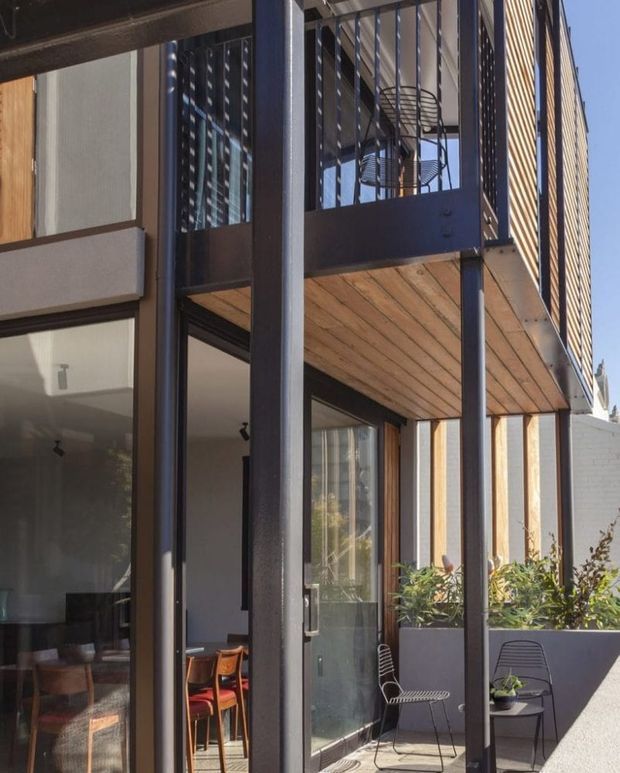 Close-up exterior view of a contemporary multi-level residential facade with dark steel structural framing, extensive glazing, timber-lined balcony soffits, vertical screened balcony elements, and a small planted terrace. Interior dining and living areas are visible through the glass.