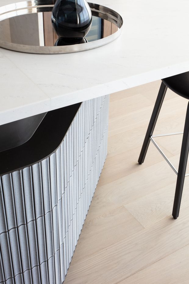 Close-up detail of a contemporary kitchen island or breakfast bar. The island has a white veined stone benchtop, a vertically fluted light-grey feature base, and a contrasting black inset or curved seating edge. A black bar stool and a reflective round tray with a dark decorative vessel sit on and beside the island. Pale timber flooring completes the bright, refined interior.