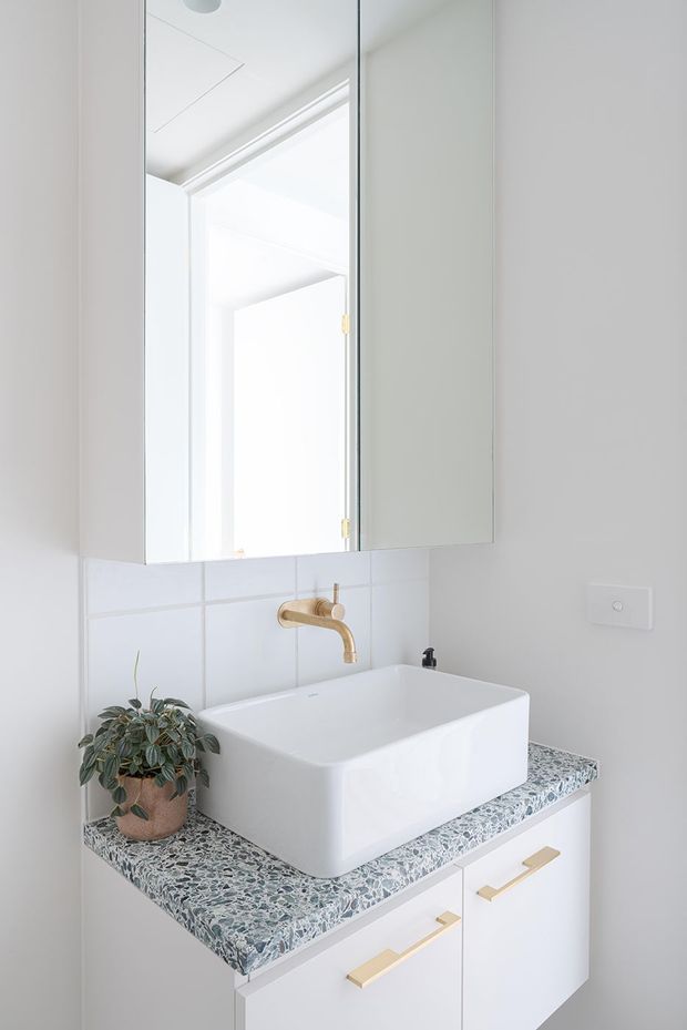 Bright, compact contemporary bathroom vanity with a white vessel basin on a terrazzo-patterned benchtop. The wall-mounted white vanity has brass/gold handles and a matching brass/gold wall-mounted tap, beneath a large mirrored cabinet. Pale tiled walls, white fixtures, and a small potted plant create a clean, refined look.