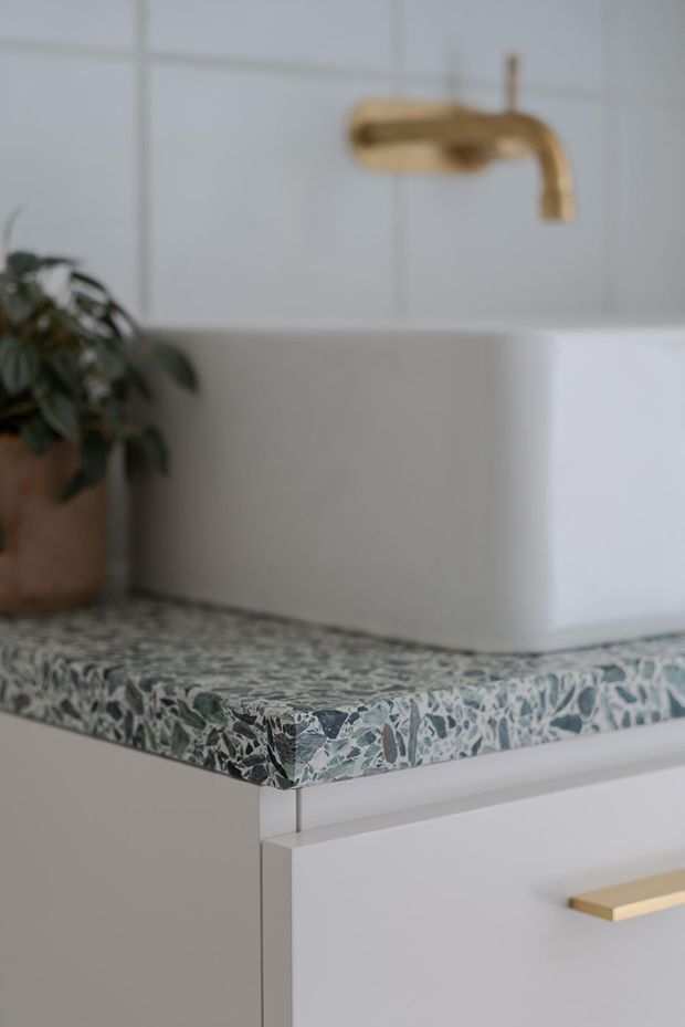 Close-up detail of a refined bathroom vanity featuring a white vessel basin, brushed brass wall-mounted tap, white tiled wall, and a patterned green-and-white terrazzo benchtop above white cabinetry with a brass handle. A potted plant adds an organic accent.