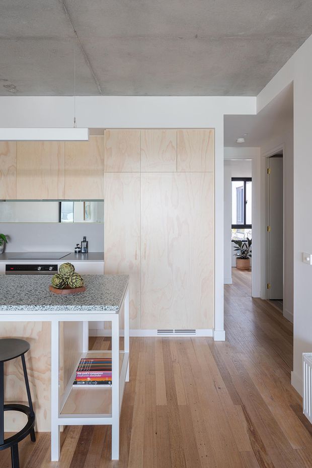 A compact, light-filled contemporary kitchen viewed from the living circulation area. Pale plywood cabinetry forms a tall bank of integrated storage and a low kitchen run, while a speckled grey stone island with white supports occupies the foreground. The space has warm timber flooring, white walls, a raw concrete ceiling, minimalist linear lighting, and a hallway leading toward a bright adjoining room.