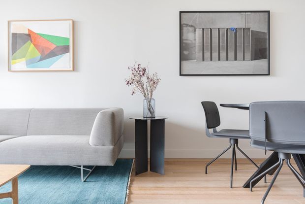 A bright, refined contemporary living and dining area with a light grey sofa, turquoise area rug, pale timber flooring, black side table, sculptural dried floral arrangement, framed abstract artwork, and a compact dining table with modern grey chairs. The room has white walls, simple furniture, and a calm, curated aesthetic.