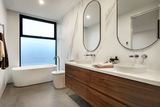 Contemporary bathroom featuring a freestanding white bathtub beneath a black-framed frosted window, a double floating timber vanity with white integrated basins, wall-mounted chrome tapware, large rounded mirrors, marble-look wall cladding, and grey floor tiles. The room is bright, refined, and minimally styled.