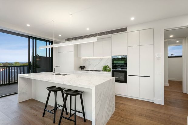 Bright contemporary kitchen with a large white marble-look island and waterfall edge, two black bar stools, integrated white cabinetry, built-in black appliances, and a subtle marble backsplash. Warm timber flooring contrasts with the white joinery. Floor-to-ceiling glazing opens to a balcony or terrace with a distant landscape view, while a hallway is visible beyond the kitchen.