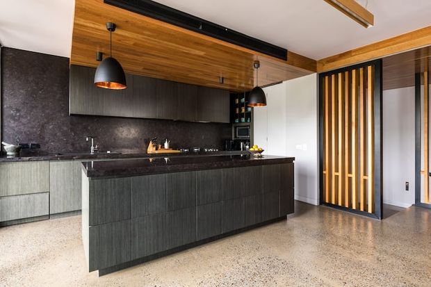 Contemporary open-plan kitchen dominated by a large dark island with a stone benchtop, dark vertical-grain cabinetry, and matching dark splashback. The room features a warm timber-lined ceiling, black pendant lights, integrated appliances, and a timber-and-black slatted screen separating the kitchen from the adjacent area. A polished speckled floor and light walls balance the darker joinery.