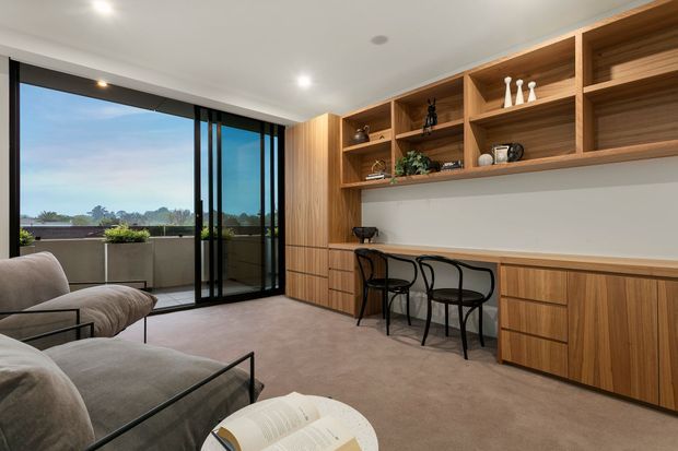 A refined contemporary living room with a built-in timber study and display wall, including open shelving, drawers, and a long desk surface with three black chairs. A soft neutral sofa and coffee table occupy the foreground, while full-height black-framed sliding glass doors open to a balcony with distant landscape views. The room has recessed ceiling lights, pale walls, and warm timber joinery.