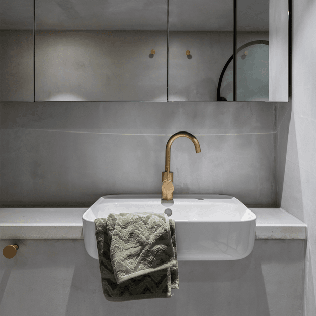 Close-up view of a compact contemporary bathroom vanity with a white wall-mounted basin, brushed brass gooseneck tap, grey microcement-style wall and cabinetry surfaces, and mirrored storage above. A textured grey towel hangs from the basin front.