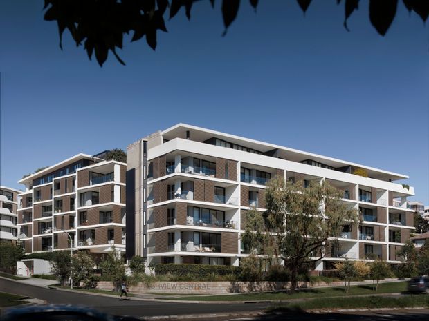 Exterior view of a contemporary multi-storey apartment complex with several connected residential blocks, projecting balconies, extensive glazing, white structural framing, and dark brick or panelled facade sections. The building sits within landscaped grounds along a suburban street under clear daylight.