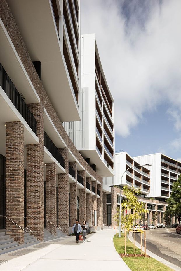 Street-level view of a contemporary multi-storey apartment or mixed-use development. The facade combines exposed red-brown brick columns and walls with white projecting floor slabs, dark metal balcony balustrades, and vertical white cladding panels. Repetitive balconies and recessed glazing line the upper levels, with a broad pedestrian sidewalk, young street trees, and a curved streetlight in the foreground.
