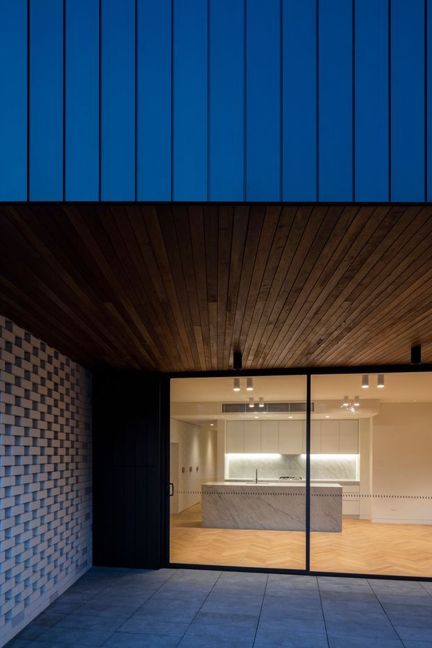 Exterior view looking through large black-framed glazing into a contemporary kitchen. The covered outdoor area has a dark timber-lined soffit, blue-toned vertical facade panels above, a textured grey brick wall to the left, and large grey tiled paving. Inside, a bright kitchen features white cabinetry, a pale stone island, patterned light timber flooring, and integrated lighting.