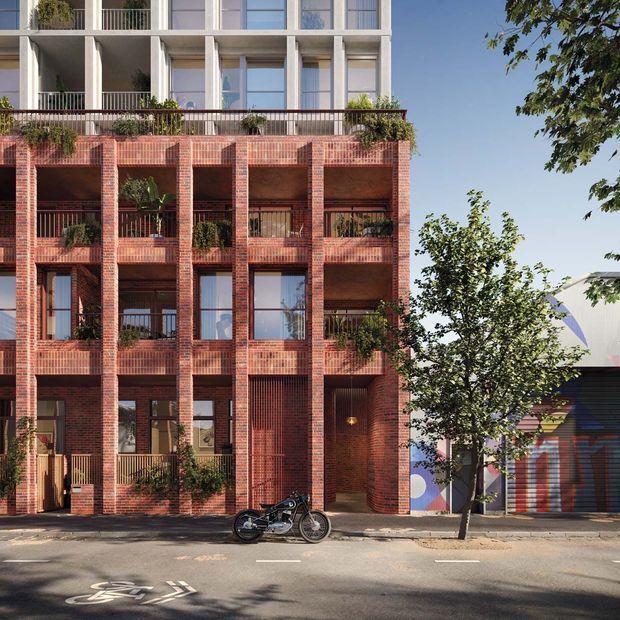 Street-facing multi-storey residential apartment building with a distinctive red brick facade, recessed balconies, dark metal balustrades, large glazed openings, and a lighter upper level with projecting balconies. Mature street trees and a neighbouring colourful garage frontage are visible in the urban streetscape.