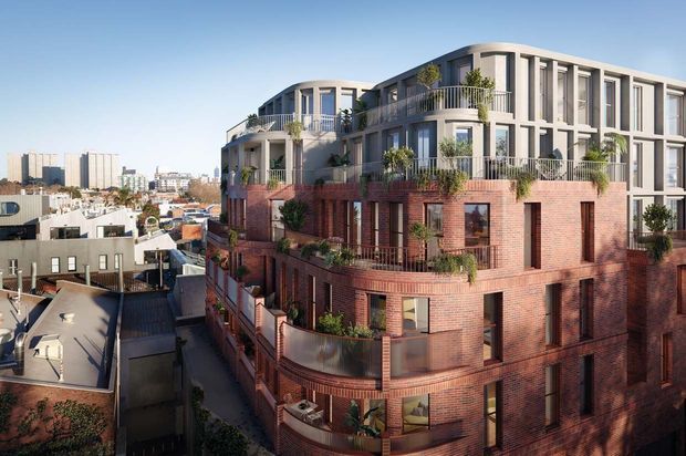 Architectural exterior rendering of a multi-storey urban residential apartment building. The building combines a red brick lower and middle facade with a light-coloured upper level featuring deep vertical window reveals, curved corners, balconies, and abundant planted terraces. Dense city buildings are visible in the background.