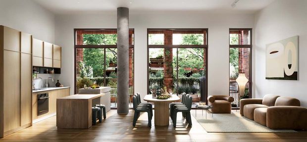 A bright, open-plan contemporary living and dining space with a compact kitchen along the left wall. Warm timber cabinetry and flooring contrast with white walls, a central concrete column, exposed red-brick window reveals, large steel-framed windows, a round dining table with dark chairs, and a relaxed brown sofa seating area.