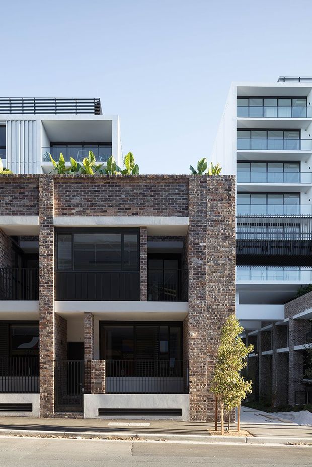 Street-facing view of a contemporary multi-storey residential development. The foreground building features exposed dark red-brown brick walls, pale rendered concrete bands, black metal balcony railings, and large dark-framed windows. A taller white apartment building with stacked balconies and extensive glazing is visible behind it, along with rooftop planting and a paved pedestrian entrance.