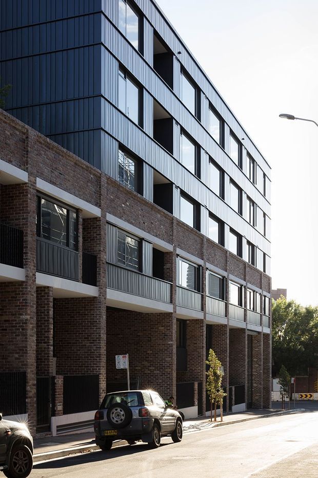 Street-level exterior view of a multi-storey contemporary residential or mixed-use building. The lower levels feature exposed dark brown brick, recessed ground-floor entries, balconies, and dark metal fencing, while the upper levels use vertically ribbed dark metal cladding with large glazed openings and projecting balcony forms. The building has a strong linear facade composition and sits within an urban streetscape.