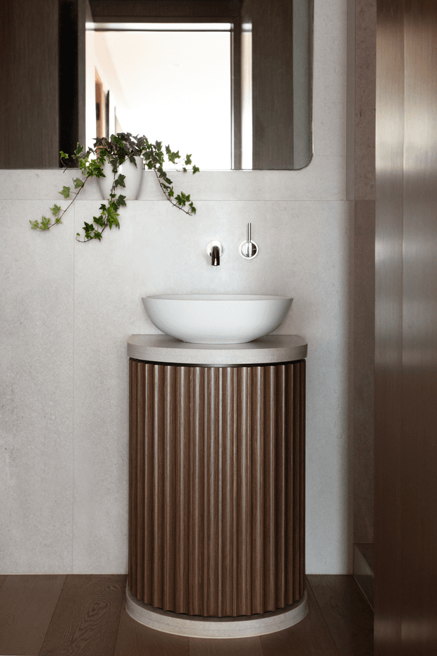 A refined bathroom vanity vignette featuring a white vessel basin on a cylindrical fluted dark timber vanity with a pale stone benchtop. The wall is clad in large light-grey stone-look panels, complemented by brushed metallic tapware, a rounded-edge mirror, dark timber flooring, and a trailing green plant. The overall composition is warm, contemporary, and sculptural.