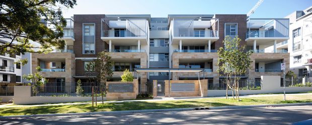 Street-facing view of a contemporary multi-storey apartment building with a symmetrical facade, exposed warm-toned masonry or stone-look wall sections, white rendered elements, extensive balconies with vertical metal balustrades, recessed parking or entry bays, and landscaped frontage. Neighboring apartment buildings and a construction crane are visible in the background.