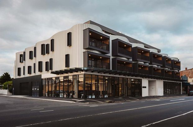 Street-level exterior photograph of a contemporary multi-storey mixed-use building. The facade combines a white vertically ribbed upper volume with dark projecting balconies and cladding, extensive ground-floor glazing, and a covered pedestrian canopy. Urban streetscape setting under an overcast sky.