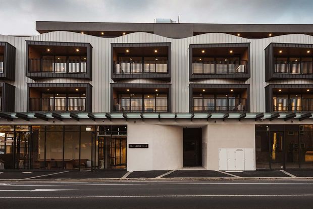 Street-facing view of a contemporary multi-storey commercial or residential building with a repetitive facade of projecting upper-level balconies, dark metal railings, vertical light-coloured cladding, deep brown soffits, and extensive ground-floor glazing. A paved urban street and entry forecourt are visible in front.