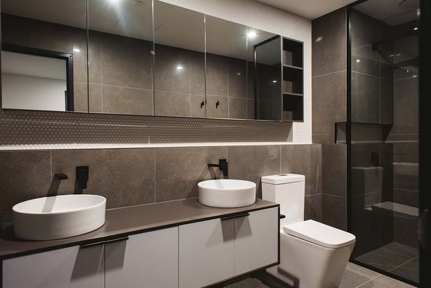 Contemporary bathroom with a double vanity featuring white vessel basins, dark engineered-looking benchtop, white wall-mounted cabinetry, a wall-hung toilet, and a black-framed glass shower enclosure. Large mirrored cabinetry and dark grey tiled walls are complemented by a small-format textured tile feature band.