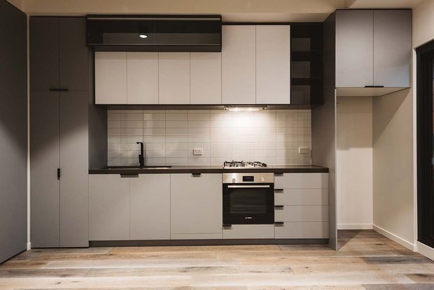 A compact contemporary galley-style kitchen with full-height dark cabinetry, pale handleless overhead and base cabinets, a tiled splashback, integrated oven and cooktop, black sink tap, and under-cabinet lighting. Timber-look flooring extends into the adjacent open area, with a tall pantry or storage cabinet at left.