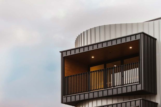 Close-up exterior view of a contemporary multi-storey building featuring a projecting balcony or recessed outdoor room. The facade combines dark vertical cladding, pale ribbed curved metal cladding, timber-lined soffits, recessed downlights, and black vertical metal balustrades.