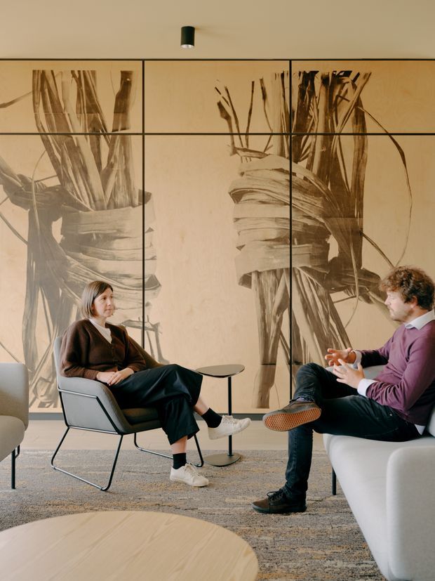 A refined contemporary lounge or meeting area with two people seated in modern armchairs around small round side tables. The room features a large sepia-toned botanical artwork or mural divided into framed panels, a neutral textured carpet, pale timber coffee table, and sculptural upholstered seating. A compact black ceiling spotlight provides focused lighting.