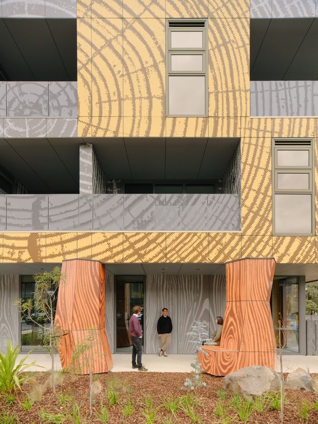 Exterior view of a contemporary multi-storey building with a striking patterned facade in warm ochre, grey, and orange tones. The facade includes projecting balconies with pale patterned balustrades, dark soffits, narrow vertical windows, and a recessed ground-floor entrance framed by sculptural orange screening elements. Landscaping and several people are visible in front of the entry.