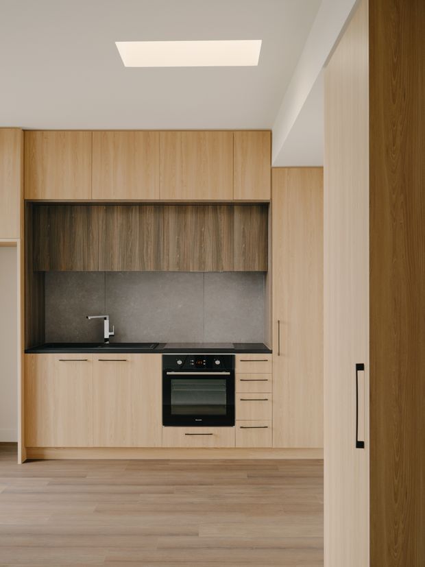A compact modern kitchen with full-height light timber cabinetry, a dark stone-look splashback and benchtop, integrated black oven and cooktop, stainless tap, and warm timber flooring. The space is viewed through a partially open doorway, with a simple recessed ceiling light and clean linear joinery.