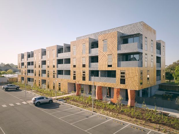 Exterior view of a contemporary multi-storey apartment building with a distinctive patterned timber-toned facade, recessed balconies, large windows, and landscaped frontage. The building overlooks a marked parking area and appears set in a suburban or campus-like environment.