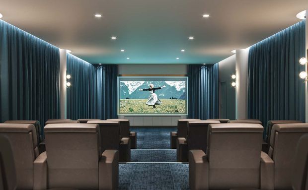 A compact private screening room with rows of upholstered recliner-style seats facing a large projection screen. Dark blue acoustic curtains line both side walls, with recessed ceiling lights and wall sconces creating a refined cinematic atmosphere. The screen displays a scenic image, and the room has stepped flooring with dark carpet.