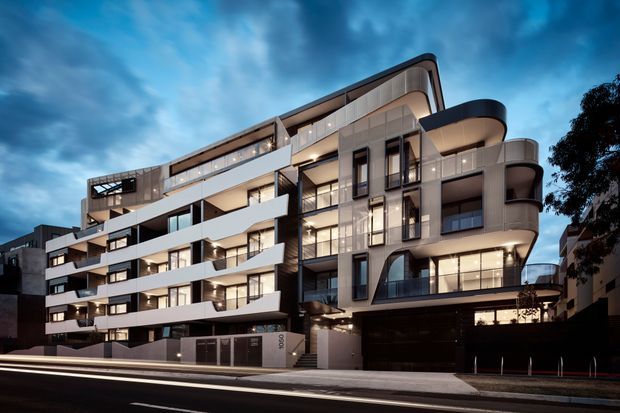 Dusk exterior photograph of a contemporary multi-storey apartment building on an urban street. The facade combines curved and angular forms, extensive glazing, projecting balconies, dark metal elements, pale rendered wall surfaces, and a dark recessed vehicle entry at street level. Warm interior lighting contrasts with the cool blue sky.