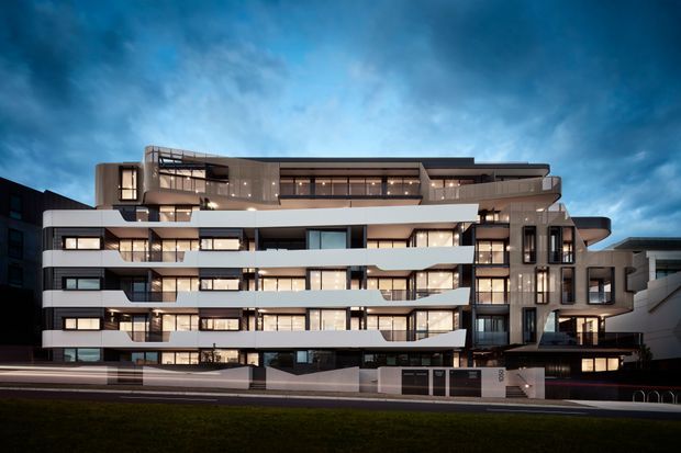 Exterior architectural photograph of a contemporary multi-storey apartment building at dusk. The facade features layered horizontal balconies, extensive glazing, dark vertical accents, warm interior lighting, and a sculptural upper-level roofline. A street frontage and entry boundary are visible in the foreground.