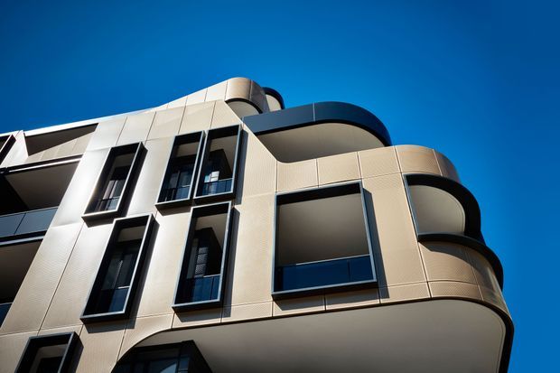 Upward-looking exterior photograph of a contemporary multi-storey apartment building. The facade combines pale beige panel cladding with dark-framed windows and strongly rounded projecting balconies, set against a clear blue sky.