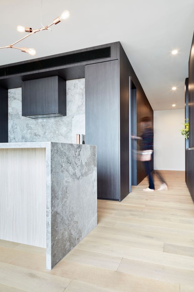 A contemporary open-plan kitchen and hallway with dark charcoal-grey integrated cabinetry, a large pale grey stone island and matching stone splashback, and light timber flooring. The space has a clean white ceiling with recessed downlights, a sculptural brass-toned multi-arm pendant, and a long linear ceiling air vent. A blurred person is moving through the hallway beside dark timber wall panelling and door openings.